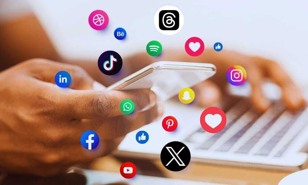 La imagen muestra a una persona sosteniendo un teléfono móvil mientras usa una computadora portátil en el fondo. Alrededor del teléfono flotan varios íconos de redes sociales y aplicaciones populares como TikTok, Instagram, Facebook, WhatsApp, Threads, X (anteriormente Twitter), LinkedIn, Spotify, YouTube, Snapchat y Pinterest, simbolizando la conexión con múltiples plataformas digitales y de redes sociales.