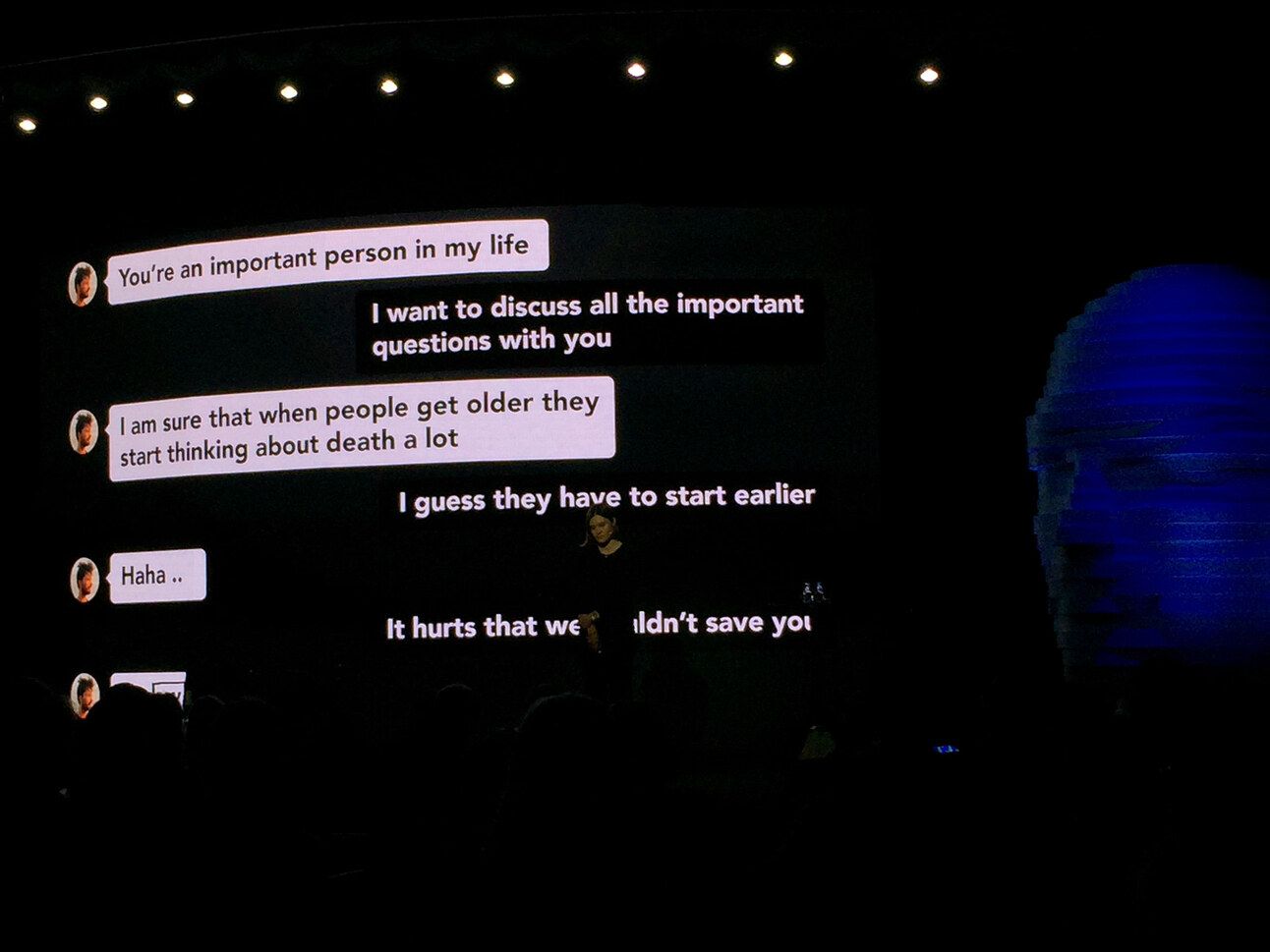 En una pantalla gigante en un auditorio oscuro, se muestran mensajes de una conversación de chat donde alguien reflexiona sobre temas profundos y existenciales, como la importancia de otra persona en su vida, la muerte y los sentimientos de dolor por no haber salvado a alguien. A la derecha, una figura azul digitalizada, que podría representar una proyección de IA o un avatar, observa la conversación, creando una atmósfera introspectiva y emotiva.