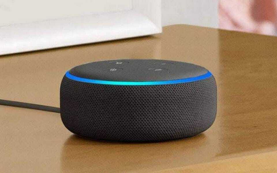 Primer plano de un altavoz inteligente Amazon Echo Dot de color negro, sobre una superficie de madera. El dispositivo muestra una luz azul alrededor de su borde superior, indicando que está activado y listo para recibir comandos de voz.